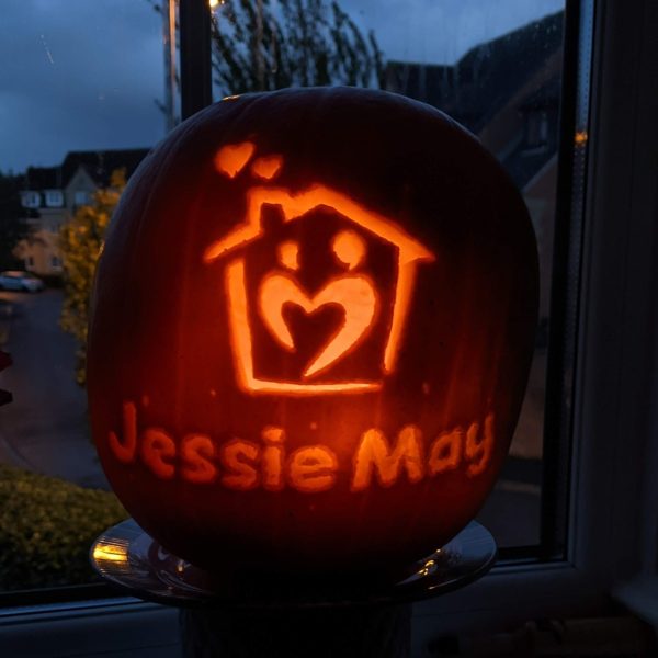 Pumpkin sitting in a dark window with light shining through showing a carving of the Jessie May house logo reading 'Jessie May' underneath