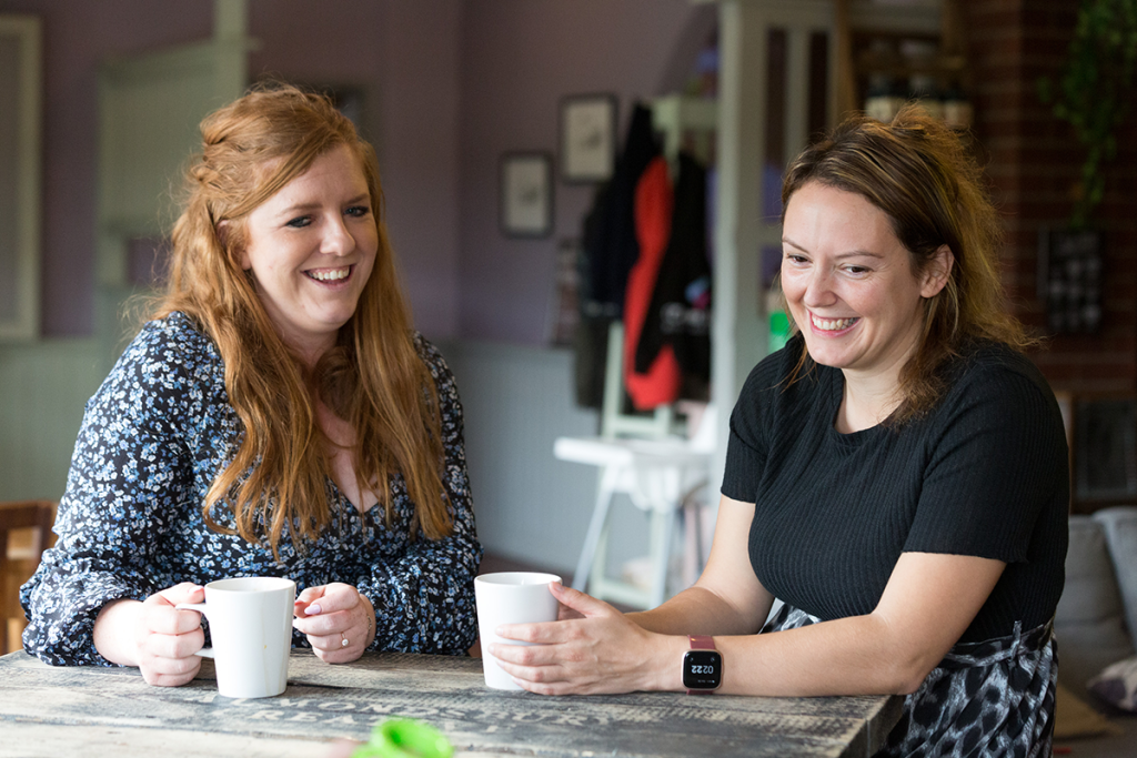 Two women, Mum's Beccy and Hayley, sat together laughing and drinking coffee