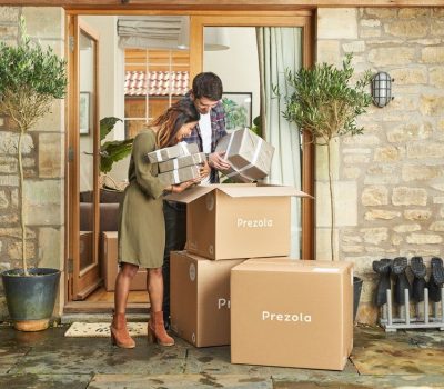 Woman and man stood outside a house looking through a pile of boxes with the word 'Prezola' written on them