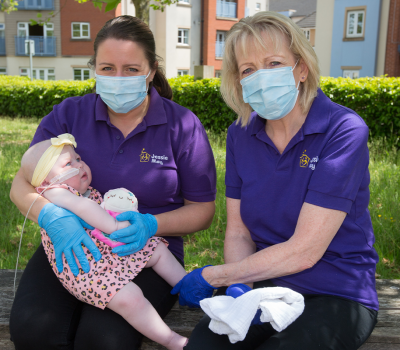 Two Jessie May nurses, female, wearing purple polo shirts, surgical masks and gloves holding a young child, girl, with a nasal-tube
